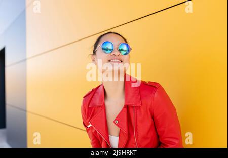 Femme souriante portant des lunettes de soleil debout devant le mur jaune Banque D'Images