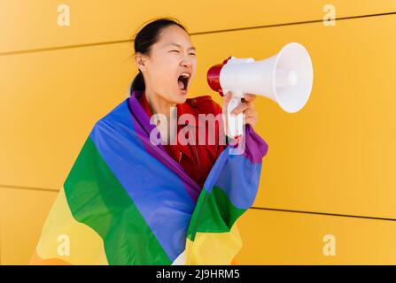 Femme enveloppée dans un drapeau arc-en-ciel criant à travers le mégaphone par le mur jaune Banque D'Images
