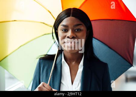 Femme d'affaires souriante tenant un parapluie coloré dans le bureau Banque D'Images