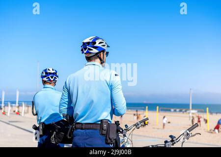 Deux policiers patrouillent sur la promenade du bord de mer à vélo. Les gens brûlent le soleil sur la plage publique de la ville, sur la côte Atlantique. Patrouille de police à vélo le Banque D'Images