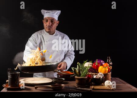 Chef masculin en uniforme blanc tenant une poêle à frire, sautant des spaghetti avec des légumes frais volant dans l'air avant de servir dans un restaurant Banque D'Images