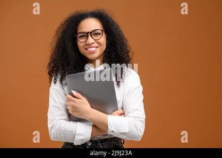 Jeune femme afro-américaine avec une coiffure afro portant une tenue élégante et décontractée et des lunettes de vue stylées isolées sur marron, portant un ordinateur portable, souriant, employée de bureau, étudiante Banque D'Images