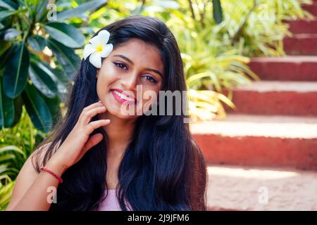 Gros plan portrait tête de l'attitude confiant sourire heureux jolie jeune femme fond d'arbres flous, fleurs de plumeria. Émotion humaine positive Banque D'Images