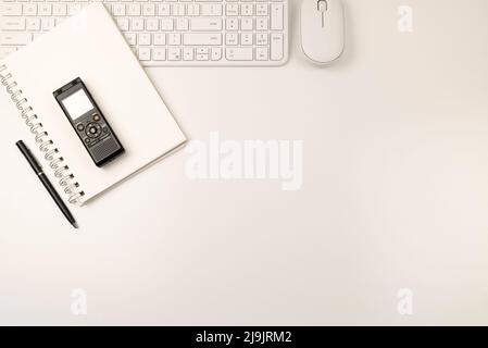 Clavier, souris et bloc-notes blancs sur le côté gauche avec stylo noir et enregistreur vocal. La graisse repose sur un arrière-plan blanc avec un espace de copie. Banque D'Images