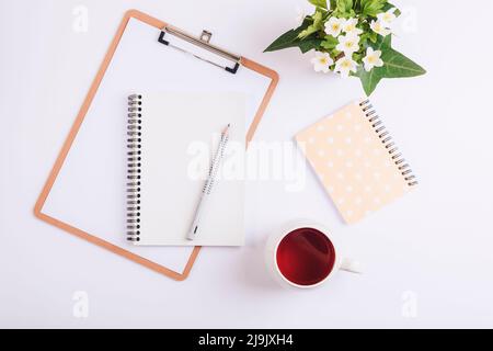 Bureau blanc avec presse-papiers, tasse de thé, crayon, carnet et bouquet de fleurs printanières. Flat lay, vue de dessus, maquette. Banque D'Images