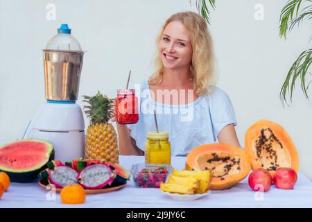 Style de vie blonde femme mélangeant jus de mixeur bocaux en verre avec smoothies et paille dans le balcon d'été.fruits exotiques tropicaux sur la table.nettoyage Banque D'Images