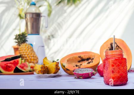 Fruits tropicaux Ingrédients grenade, ananas, papaye, fruit dragon, orange et pastèque avec mélangeur sur table blanche fond d'été feuille de palmier Banque D'Images