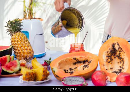 Style de vie blonde femme mélangeant jus de mixeur bocaux en verre avec smoothies et paille dans le balcon d'été.fruits exotiques tropicaux sur la table.nettoyage Banque D'Images