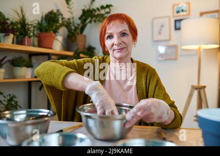 Femme sénior aux cheveux rouges en gants de protection plantant des plantes dans des pots pour les semis tout en étant assise à la table à la maison Banque D'Images