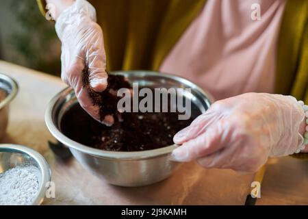 Gros plan du jardinier en gants de protection fertilisant le sol en pot pour planter des semis tout en étant assis à la table Banque D'Images