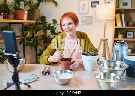 Femme senior faisant son blog sur le jardinage, elle s'asseyant à table et transplantant des fleurs dans d'autres pots n caméra de smartphone Banque D'Images