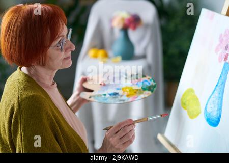 Femme sénior à cheveux rouges en lunettes de vue regardant sa photo sur toile pendant la peinture en classe Banque D'Images