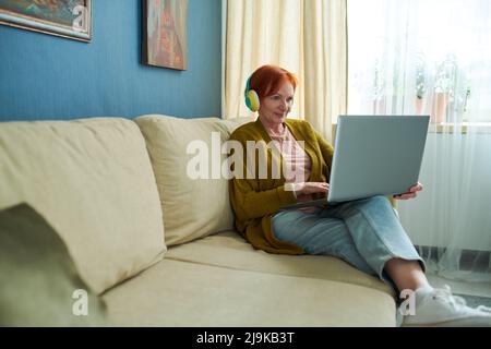 Femme âgée dans un casque sans fil utilisant un ordinateur portable tout en se reposant sur un canapé dans le salon, elle ayant la consultation en ligne Banque D'Images