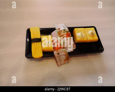 Tamagoyaki à la feuille d'or ou à l'œuf de sushi sucré avec un rouleau de sushi au bacon dans une assiette carrée noire sur une table brune, cuisine fusion japonaise Banque D'Images