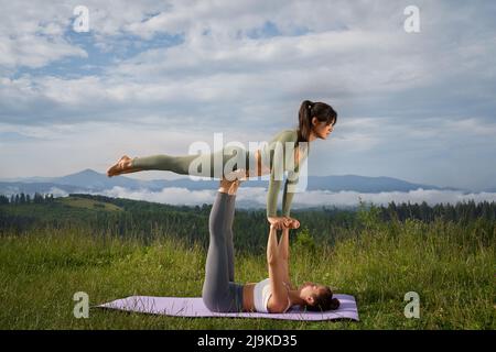 Des femmes sportives en vêtements d'activité faisant du yoga acroo parmi les belles montagnes d'été. Deux femmes caucasiennes s'entraîner régulièrement ensemble à l'air frais. Banque D'Images