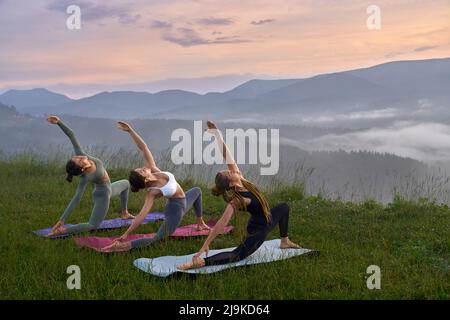 Jeunes femmes sportives en vêtements d'activité faisant des exercices de yoga parmi les montagnes vertes. Des femelles athlétiques s'étirent sur un tapis de yoga pendant le coucher du soleil d'été. Banque D'Images