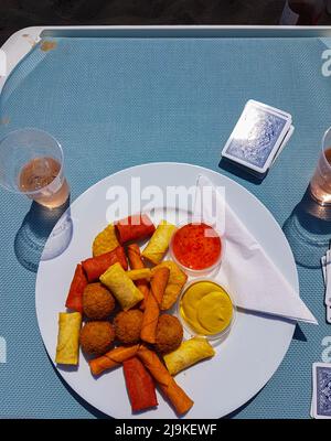 Délicieux en-cas variés sur une assiette qui est sur un lit de plage avec une tasse de vin de rose avec glace et un jeu de cartes à côté Banque D'Images