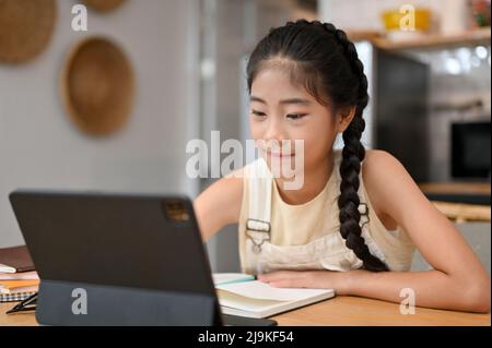 Jolie petite fille asiatique s'assoit dans une salle à manger, étudie la leçon en ligne et prend un cours en ligne pendant la pandémie. Enfants Homeschool concept. Banque D'Images