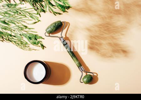 Rouleau facial et bol crème avec ombre de branche de cyprès sur fond beige clair. Cosmétiques naturels, aromathérapie, concept de soins de beauté. Vue de dessus, Banque D'Images