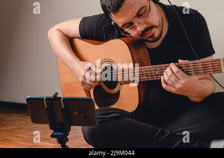 le jeune homme du caucase, avec des lunettes et en noir, à la maison assise sur le sol jouant de la guitare diffusant en direct sur les réseaux sociaux sur son téléphone. Banque D'Images