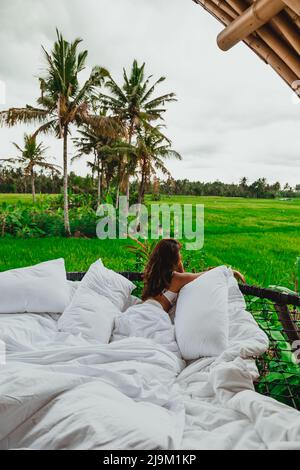 jeune femme asiatique en draps blancs donnant sur les rizières de bali et les cocotiers depuis son balcon de villa en bambou Banque D'Images