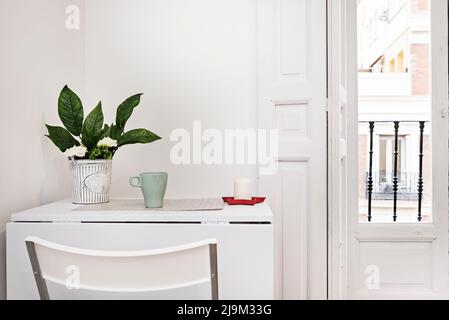 Appartement studio avec table à manger pliante blanche avec faux vase, bougie dans un cendrier rouge, balcon avec fenêtres en bois blanc vintage, volets et tapis Banque D'Images