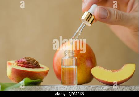 Une main femelle tient un compte-gouttes avec de l'huile essentielle de pêche. Cosmétiques naturels pour les soins de la peau. Bouteille de sérum et pêches fraîches sur podium blanc Banque D'Images