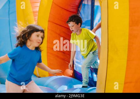 Adorable contenu petite sœur et frère avec des cheveux foncés dans les t-shirts courir à l'intérieur de trampoline gonflable coloré tout en passant du temps ensemble Banque D'Images