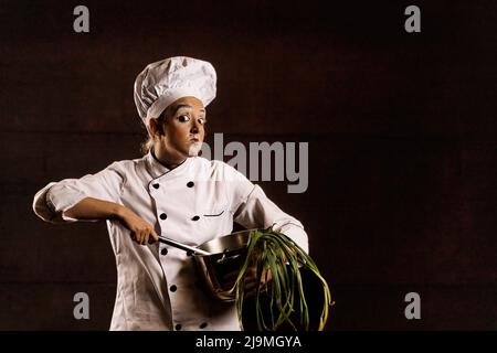 Clown jeune femme avec un maquillage brillant en uniforme de chef et chapeau regardant l'appareil photo avec curiosité tout en remuant le plat dans une casserole contre le backgroun noir Banque D'Images