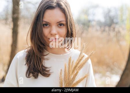 Jeune femme aux longs cheveux foncés et aux yeux bruns tenant un bouquet d'épillets de blé et regardant la caméra en campagne Banque D'Images