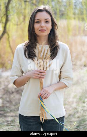 Jeune femme gaie avec de longs cheveux foncés dans des vêtements décontractés souriant et regardant l'appareil-photo tout en étant debout dans la nature avec un tas de spikettes de blé liées Banque D'Images