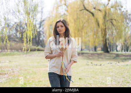 Jeune femme sérieuse avec de longs cheveux foncés dans des vêtements décontractés regardant l'appareil-photo tout en se tenant dans la nature avec un bouquet de spikettes de blé attaché avec bleu et Banque D'Images