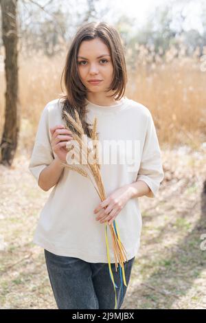 Jeune femme sérieuse avec de longs cheveux foncés dans des vêtements décontractés souriant et regardant l'appareil-photo tout en étant debout dans la nature avec un tas de spikettes de blé attaché W Banque D'Images