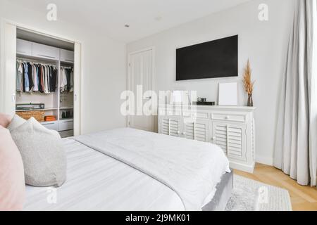 Téléviseur avec écran noir accroché au mur au-dessus de l'armoire près du lit mou dans la chambre lumineuse avec placard Banque D'Images