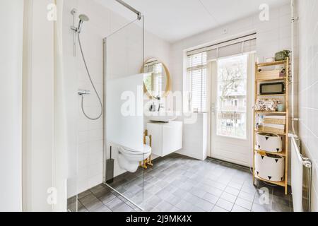 Intérieur de la salle de bains spacieuse avec cuvette de toilettes en céramique blanche près de cabine de douche avec porte en verre et lavabo près de l'étagère sur le mur Banque D'Images