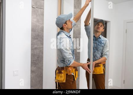 L'assembleur de mobilier employé en t-shirt blanc et combinaison-pantalon vérifie la porte coulissante en bois de l'armoire moderne avec miroir. Banque D'Images