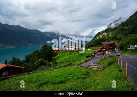 Vue sur la route de montagne avec des maisons en bois de chalet rouge le long des rives du lac de brienz entouré de montagnes alpines et de prairies verdoyantes. Banque D'Images