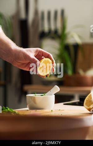 Gros plan vertical moyen d'un homme méconnaissable tenant un morceau de citron au-dessus du mortier avec des épices et de la coriandre hachée pour presser le jus Banque D'Images