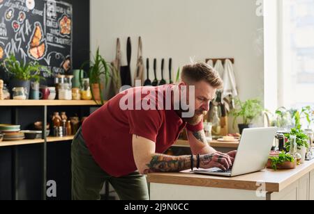 Jeune adulte barbu caucasien avec tatouages sur les armes surfer sur Internet sur ordinateur portable dans la cuisine loft à la maison Banque D'Images