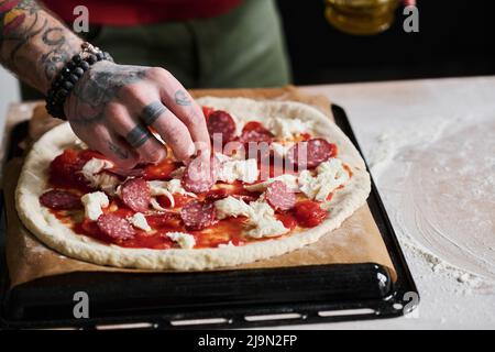 Gros plan d'un homme méconnaissable avec des bras tatoués qui cuisent une pizza à la maison en ajoutant des tranches de salami Banque D'Images