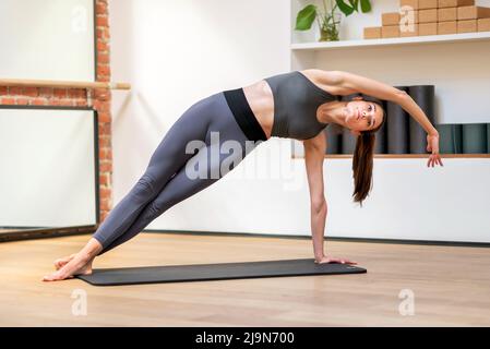 Corps complet de femme mince en forme d'activewear debout en position de planche latérale avec bras étiré tout en pratiquant le power yoga sur le tapis en studio. Salle de fitness Banque D'Images