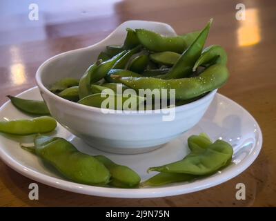 Gros plan, accent sélectif sur les gousses d'edamame dans une petite tasse blanche sur une table en bois à l'intérieur d'un restaurant Banque D'Images