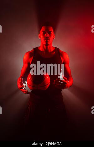 Côté joueur de basket-ball éclairé de couleur rouge tenant une balle sur fond de fumée. Sérieux homme afro-américain concentré. Banque D'Images