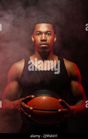 Côté joueur de basket-ball éclairé de couleur rouge tenant une balle sur fond de fumée. Sérieux homme afro-américain concentré. Banque D'Images