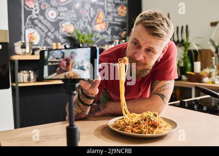 Blogueur de nourriture manger des pâtes maison tout en filmant la recette et mukbang avec smartphone sur trépied dans la cuisine Banque D'Images