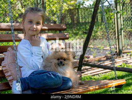 Belle petite fille est assise sur une balançoire au printemps, sourit et embrasse son petit chien. Enfants et chiens en plein air Banque D'Images