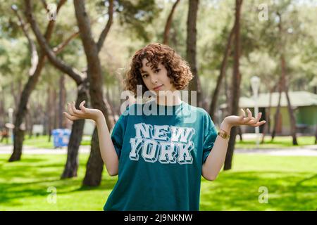 Adorable femme à tête rouge portant un t-shirt vert debout sur le parc de la ville, à l'extérieur sans indice et confus avec les bras ouverts, hausser les épaules, aucune idée et douteux f Banque D'Images
