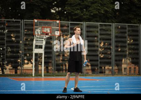 Jeune homme en chemise sueur se reposant après un entraînement intense sur la salle de sport extérieure. Beau sportif caucasien avec corps musclé dans la mode sportswear semble fatigué et épuisé. Banque D'Images