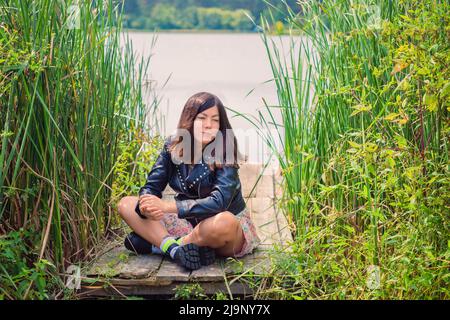 Une jolie jeune femme aux cheveux noirs est assise dans un épais d'herbe haute sur la rive de la rivière. Banque D'Images