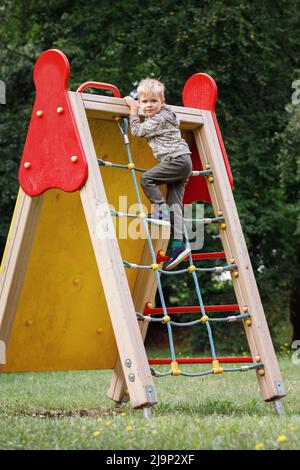 Joyeux garçon souriant jouant sur l'aire de jeux et grimpant sur le filet de corde. Concept de développement de l'enfant, de sport et d'éducation. Un enfant courageux monte à la hauteur Banque D'Images